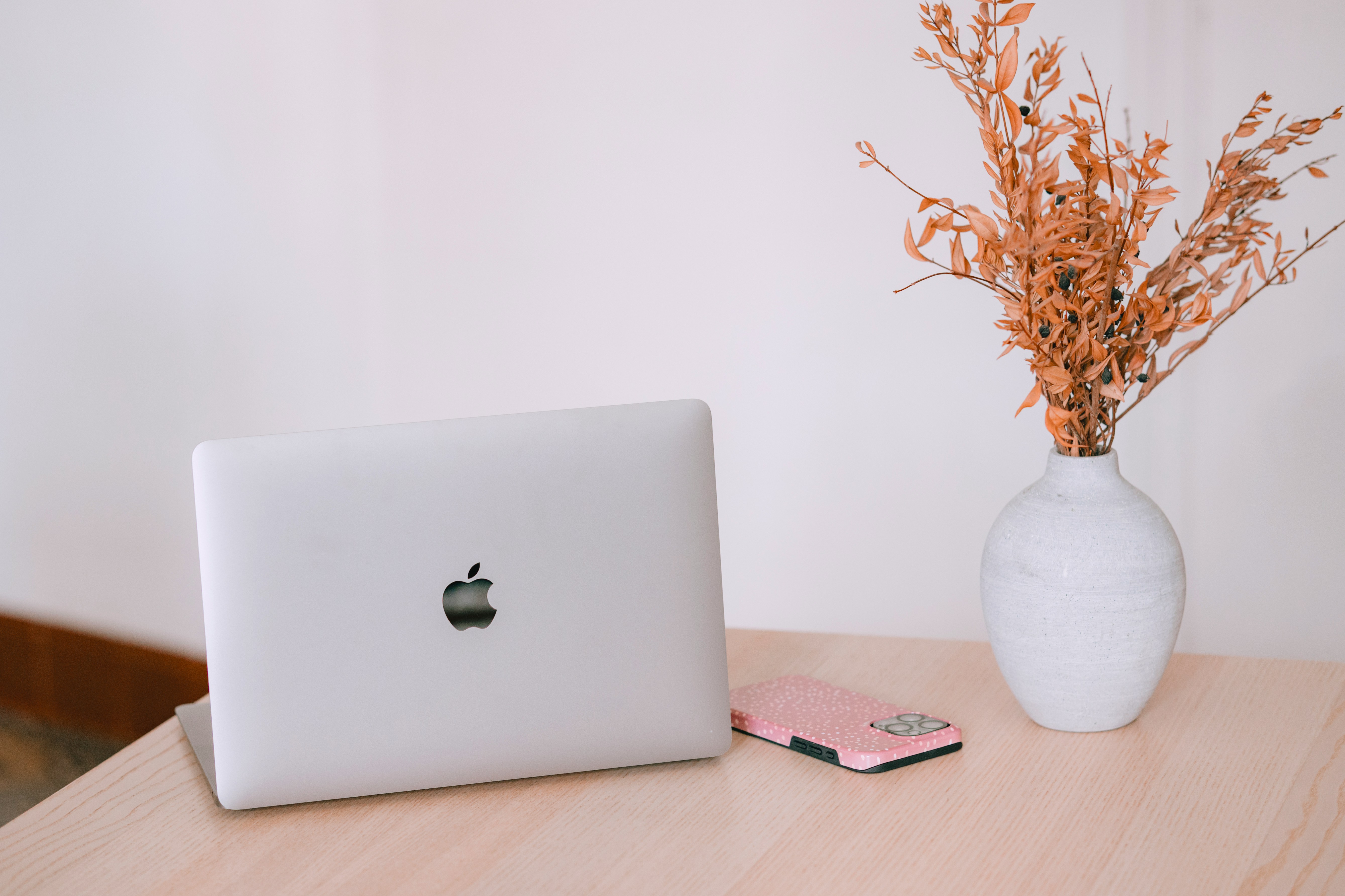 A laptop, a smartphone and a small dried flower arrangement on a wooden desk.